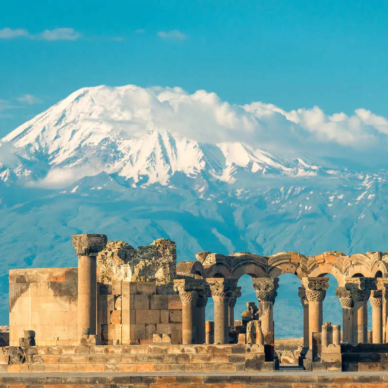 Zvartanos Temple Set Against The Backdrop Of A Snowy Mountain Range In Armenia, Caucasus Region