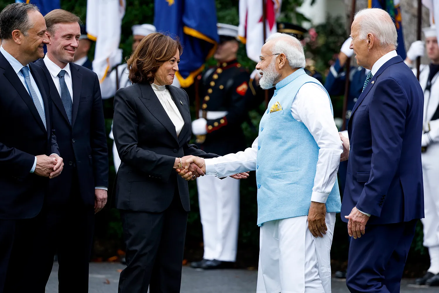 Harris (center) waring a dark suit shakes hands with Modi, wearing glasses and a blue tunic. At right is Biden in a blue suit. Emhoff and Sullivan wear dark suits behind her. All smile in greeting.