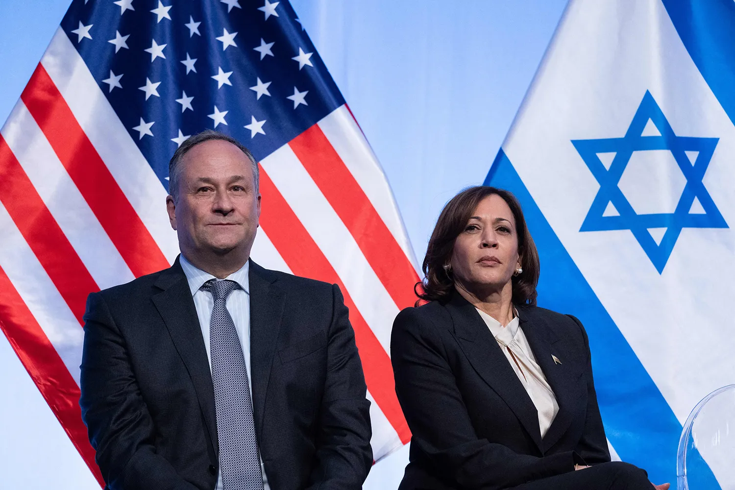 Harris and Emhoff, both wearing dark suits, look forward with serious faces as they sit in front of the flags of the United States and Israel.