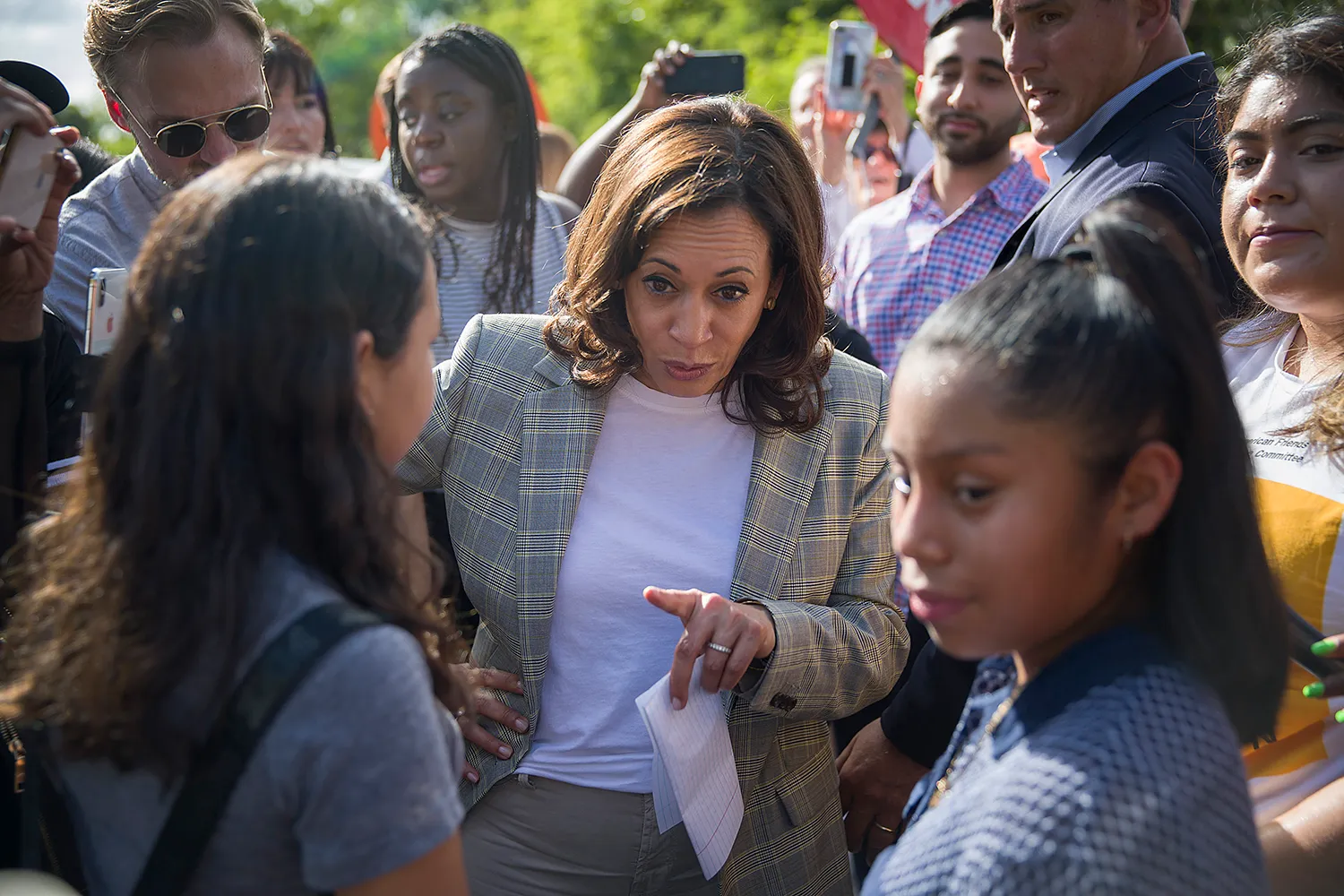 Harris, wearing a plaid suit, points as she talks with two young people in front of her. Aroound her are other people, some taking photos on their phones.