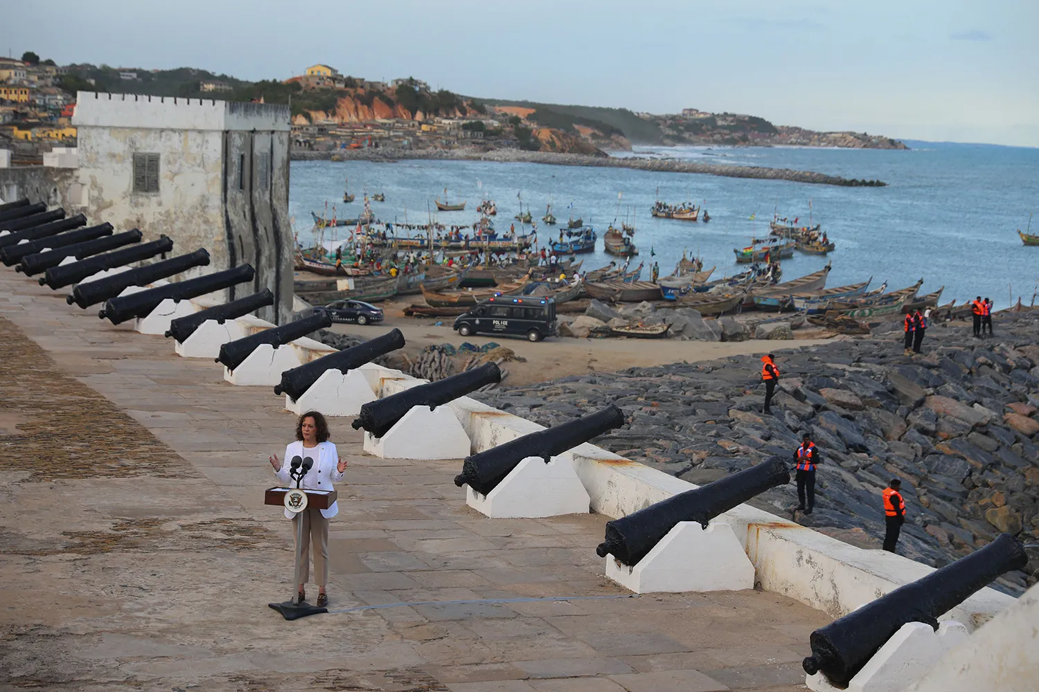 Harris stands in front of a lectern in a high-angle view that shows the grounds of a castle with cannons facing outward. Behind the wall is the rocky shore of Ghana with security officers on the rocks and small boats in the ocan in the distance.