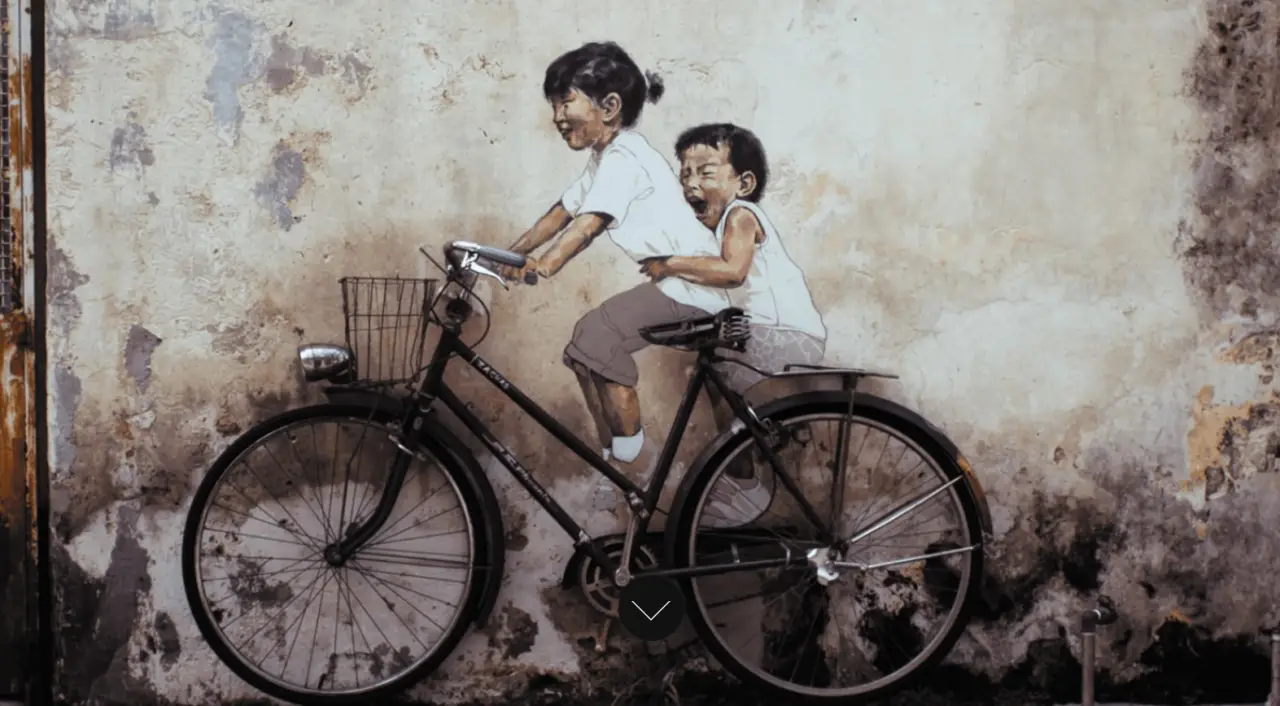 A famous piece of street art in Penang depicting two children, a girl and a boy, joyfully riding an actual bicycle that is mounted against an old, weathered wall. The mural captures a playful and nostalgic moment, blending the real bicycle with the painted figures.