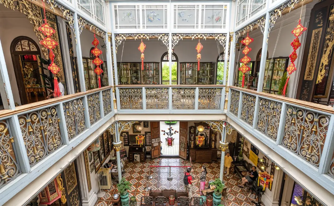 An ornate interior of a Peranakan museum featuring intricate wrought iron railings, traditional Chinese red lanterns, and decorative tiles. The museum’s multi-level atrium offers a glimpse into various rooms filled with antique furniture, showcasing the rich cultural heritage of the Peranakan community.