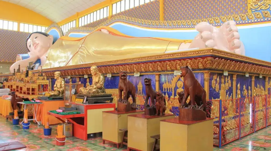 The interior of the Wat Chaiyamangalaram temple in Penang, featuring a large, golden reclining Buddha statue. The statue is surrounded by intricate carvings, smaller golden statues, and vibrant decor, with wooden animal figures positioned in the foreground.