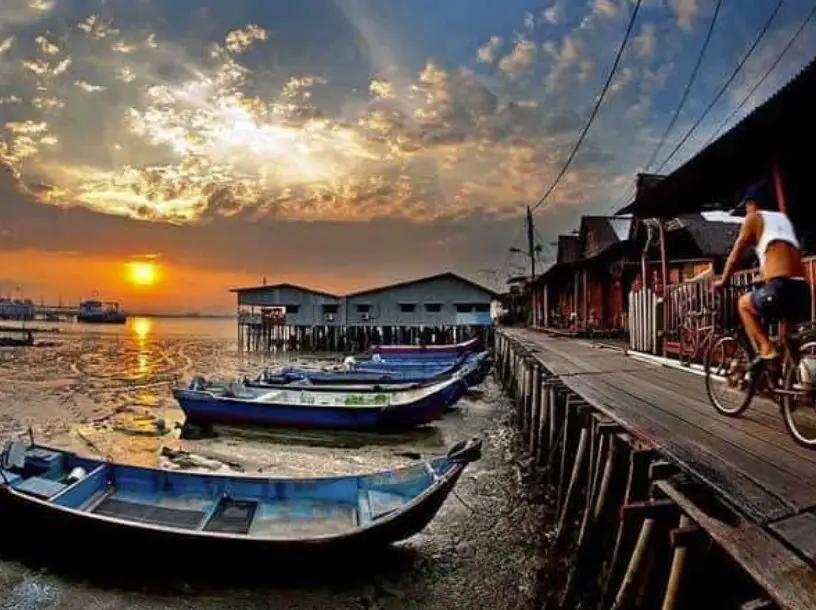 A scenic view of Chew Jetty in Penang during sunrise, showcasing traditional stilt houses over the water and small boats resting on the shore. A man on a bicycle rides along the wooden walkway that runs alongside the houses, capturing the peaceful and rustic charm of this historic waterfront community.