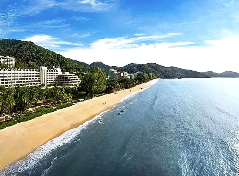 A wide view of a pristine beach in Penang, Malaysia, lined with luxurious beachfront hotels and surrounded by lush green hills. The calm blue ocean stretches out under a bright, clear sky, with gentle waves lapping at the sandy shore.