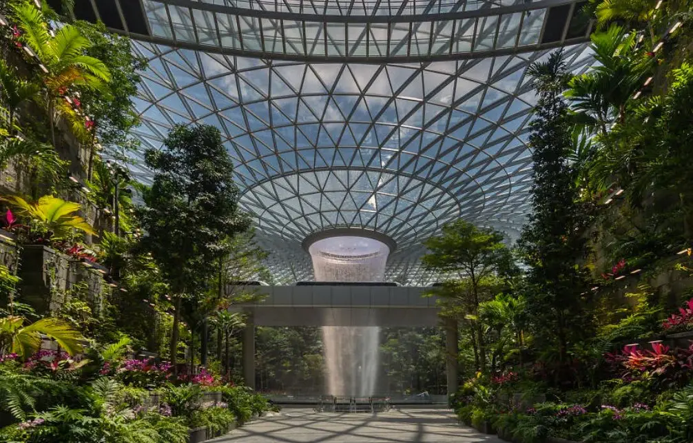 Interior view of Jewel Changi Airport showcasing the central Rain Vortex waterfall surrounded by lush greenery and a glass-domed roof.