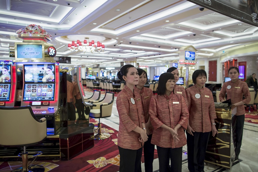 Several women in uniforms stand on the floor of a casino full of poker machines.