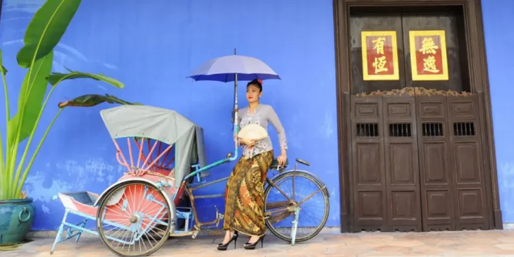 A woman dressed in traditional attire holding an umbrella, standing beside a colorful trishaw in front of the iconic blue wall of the Cheong Fatt Tze Mansion in Penang. The mansion’s wooden doors feature Chinese characters in gold and red.