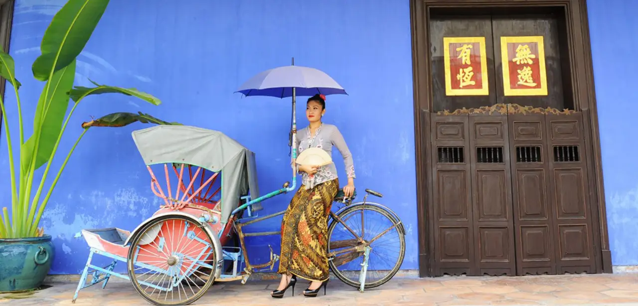 A woman dressed in traditional attire holding an umbrella, standing beside a colorful trishaw in front of the iconic blue wall of the Cheong Fatt Tze Mansion in Penang. The mansion’s wooden doors feature Chinese characters in gold and red.