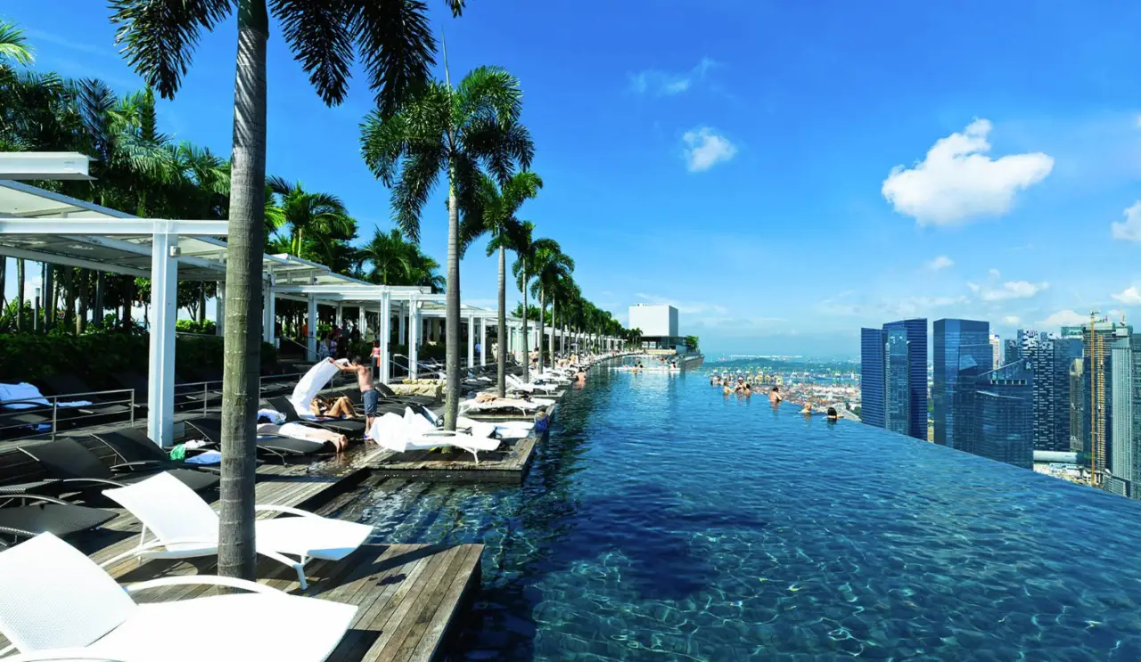 Infinity pool at Marina Bay Sands in Singapore, offering a stunning view of the city skyline and blue sky, with palm trees and lounge chairs lining the poolside.