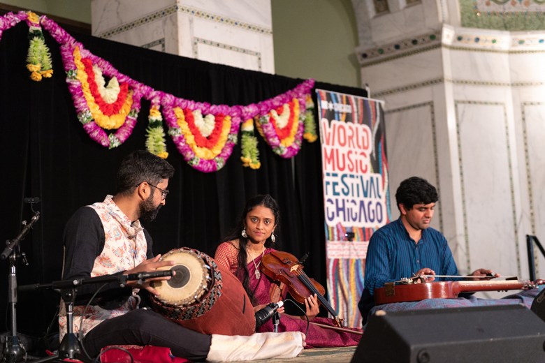 three seated Indian classical musicians at Ragamala on a low stage with a black backdrop and colorful garlands, playing mridangam, violin, and chitravina