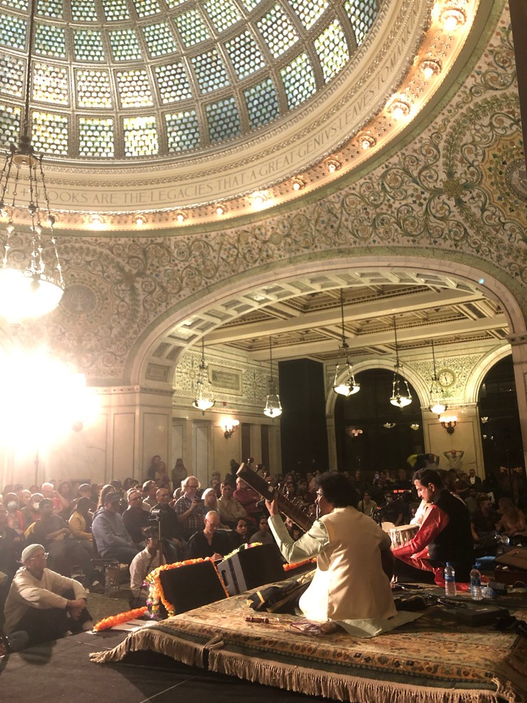 a view from behind and to the left of the Ragamala stage, where three Indian classical musicians perform, looking out at a large ornate hall with a stained-glass dome overhead and a large seated crowd